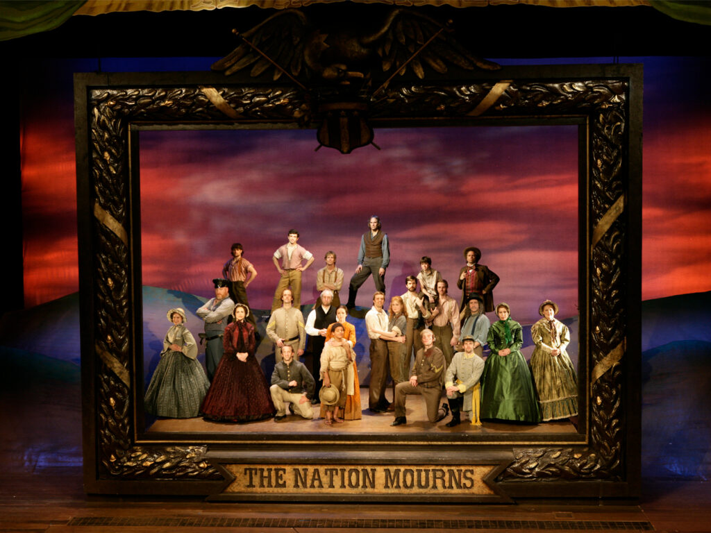 Twenty-three people stand stationary and silent, carefully posed in an 1860s photograph tableau. A gigantic gilded frame reading “A Nation Mourns” frames them.