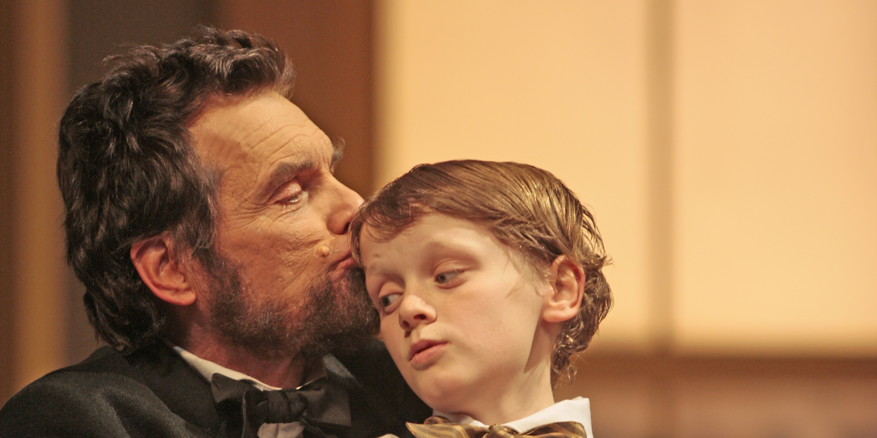 An actor portraying Abraham Lincoln holds an actor portraying his young son and kisses his forehead.