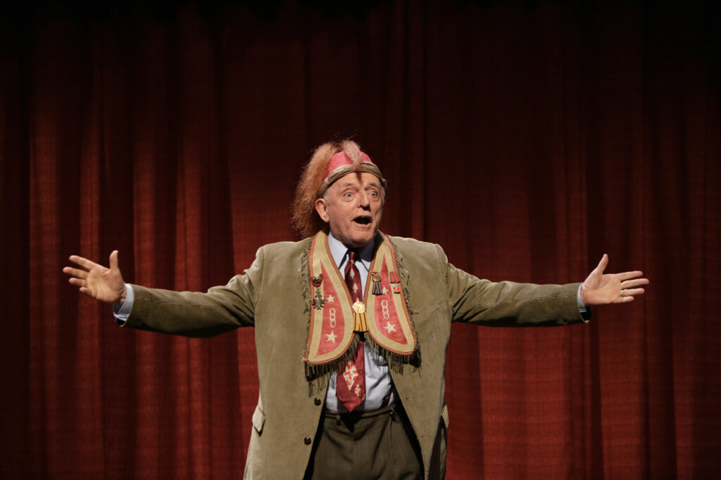 A man in a fraternal order attire of the Moose Lodge organization (red beanie-style hat with long hair tuft; red vest embroidered with gold thread and medallions) stands on a stage to introduce a play.