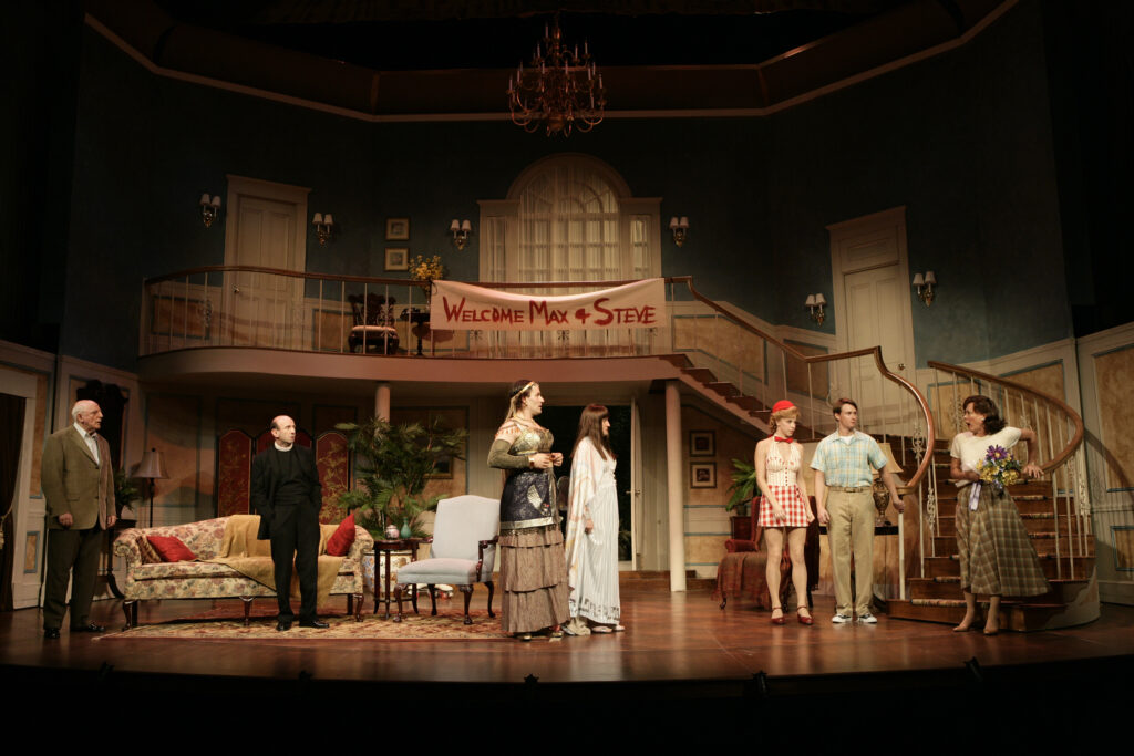 Inside the living room of a home stand a group of people with shocked faces. The groups is: an older gentleman, a priest, two male actors in ladies Shakespearean costumes, a young man in his 20s, a woman in roller-skates, and another woman who carries a large bouquet of flowers at the bottom of a large sweeping staircase. On the railing of the balcony to the second level of the house is a painted sheet reading, “Welcome Max and Steve.”