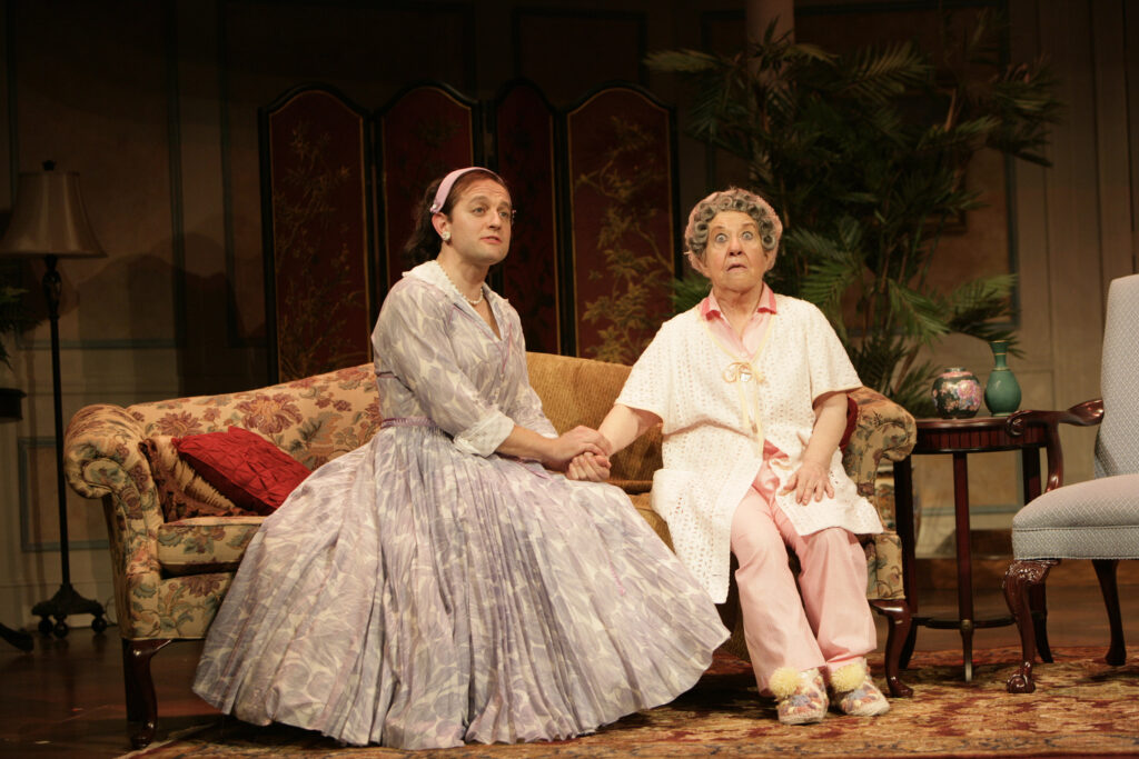 A man wearing a ladies wig and a 1950s-style dress covered in lavender tulips sits on the sofa holding the hand of an older woman. The older woman is still in her pink pajamas and bathrobe and wearing hair curlers. She has a shocked expression on her face.