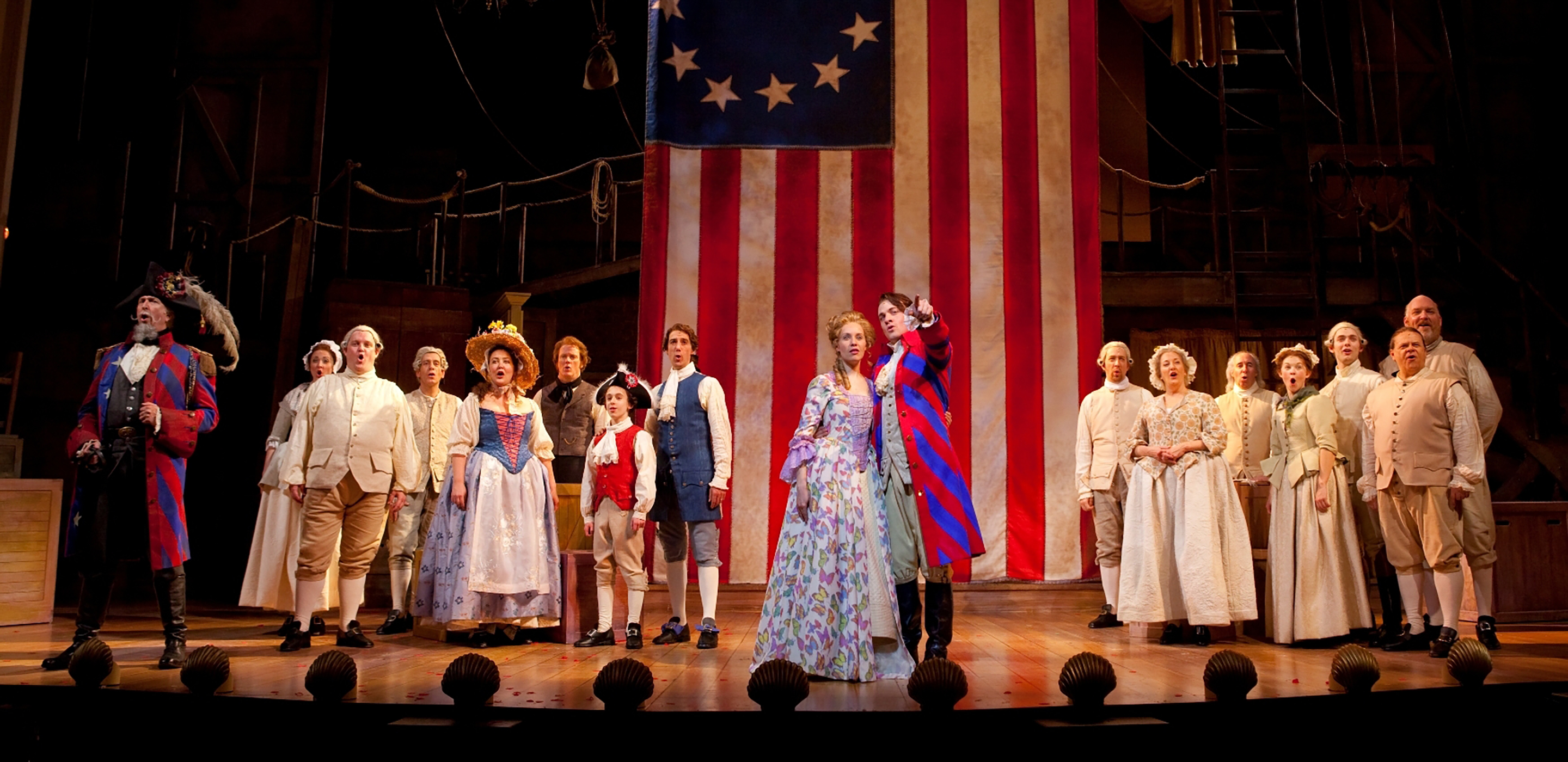 A man in a bright blue and red coat stands next to a a woman in a floral dress and points. Behind them an American flag with 13 stars hangs. To their left and right groups of people in 18th century clothing sing.