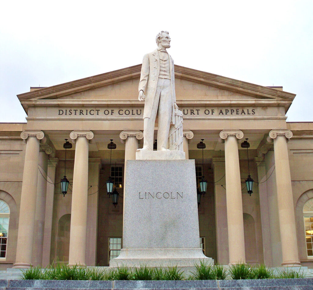 Daytime view of a statue of President Abraham Lincoln, standing and looking to the viewer’s right. Lincoln rests his hands on a bundle of sticks, bound together. A rectangular pedestal below the standing figure says “Lincoln.” Behind the statue is a light brown stone building with columns and a pediment.