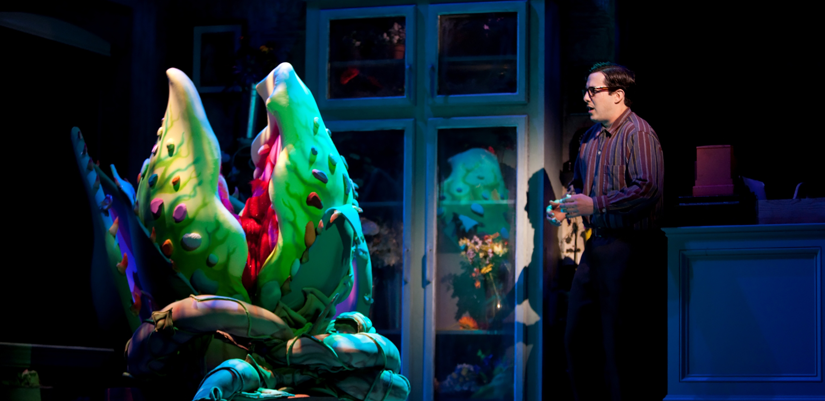 A man in glasses and a button down shirt stands in a bedroom and talks to a giant plant with a huge mouth in a pot.