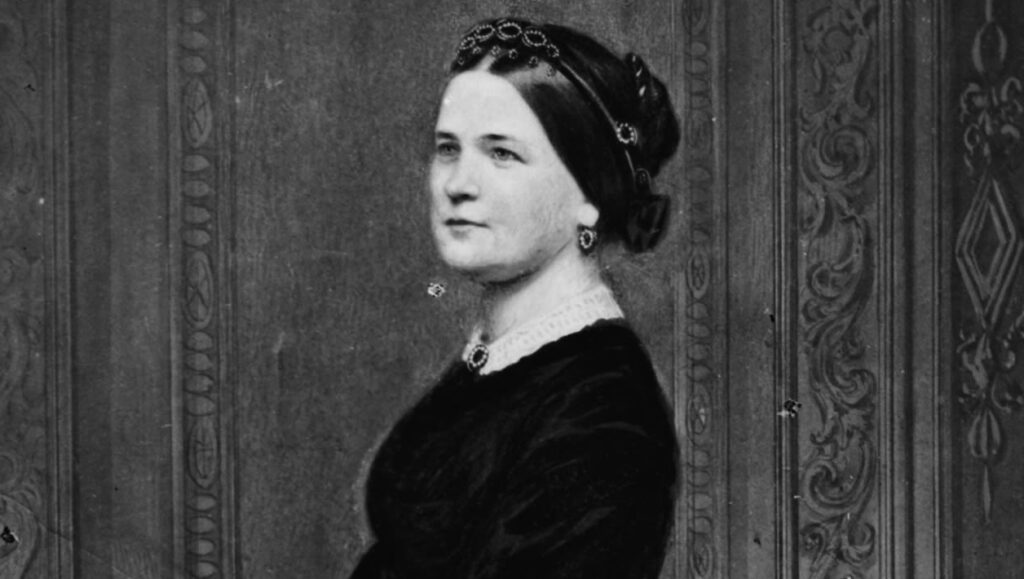 A black and white photograph of a woman in a long black dress with lace trim holding a handbag.