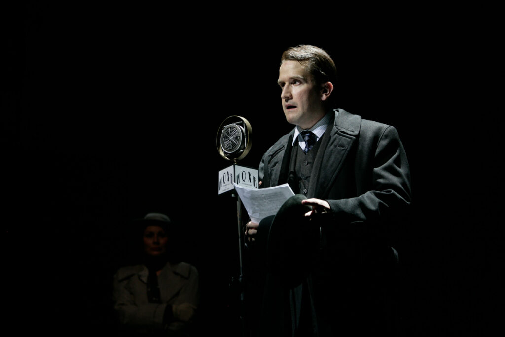 A man dressed in a three-piece suit, overcoat stands behind a radio broadcast microphone and reads from a script.