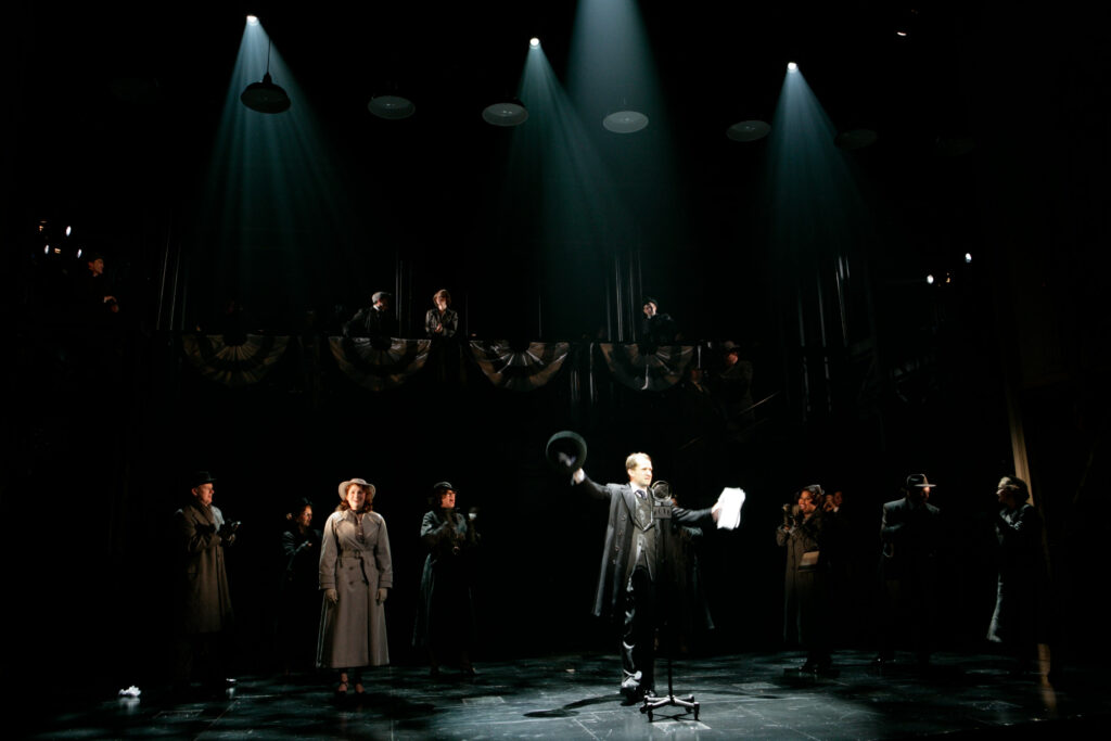 A man dressed in a three-piece suit, overcoat stands behind a radio broadcast microphone and reads from a script, while extending his hat high in his right hand. Behind him are patriotic buntings and a crowd that has gathered to listen to him speak.