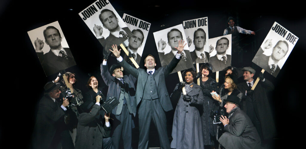 A man in a three-piece grey suit stands in the middle of a crowd with his arms raised in jubilation. The crowd carries picket signs that have photographs of his face and the words “John Doe.”