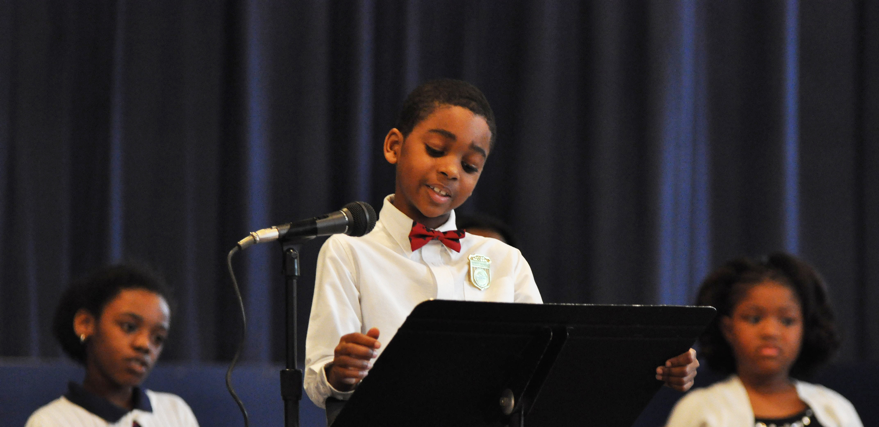 A young child wearing a white shirt and a red bowtie stands in front of a microphone and delivers a speech. Behind him two girls sit down and listen.