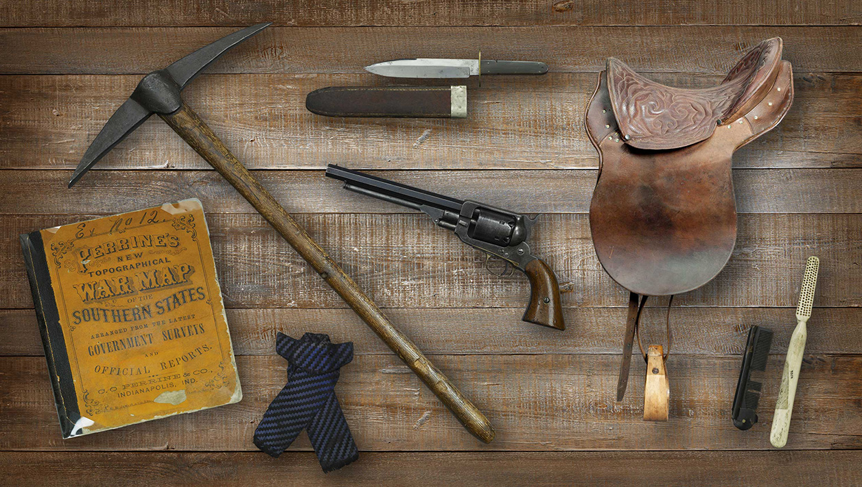 A series of objects laid out on a wooden table: A map book, a pickax, a necktie, a knife and sheath, a revolver, a saddle and comb.