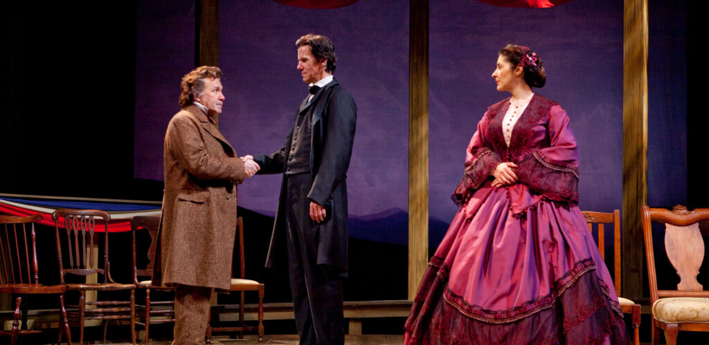 An actor portraying Stephen Douglas shakes hands with the actor portraying Abraham Lincoln. The actress portraying Adele Douglas looks on.