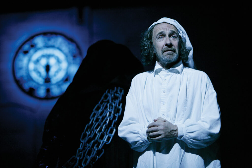 A man stands in his white nightcap and nightshirt clasping his hands. Behind him is a shadowy figure draped with heavy chain links. Farther in the distance, a large clock glows.