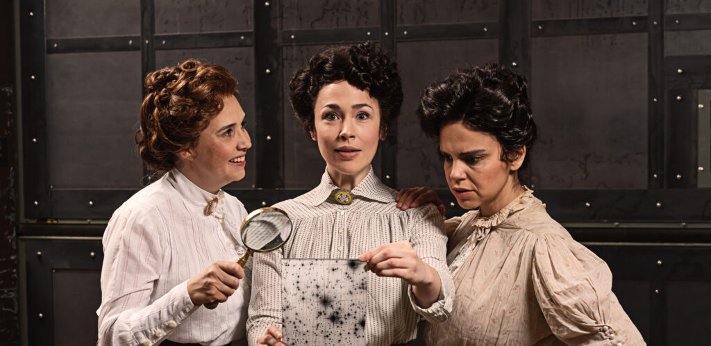 Three actresses wearing Victorian blouses and high-waisted skirts stand together. The woman in the middle holds a glass plate with a photograph of the night sky and has an expression of discovery on her face. The woman on the left holds a magnifying glass and smiles as she looks at the woman in the middle. The woman on the right stares at the details on the glass plate.