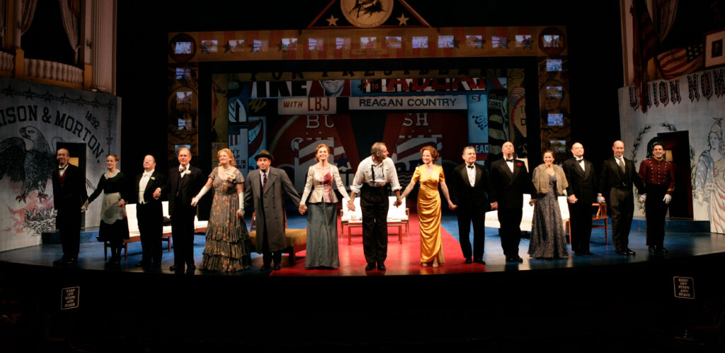 A group of actors in various 50's style clothing stand hand in hand at the edge of a stage. Behind them the stage is decorated with political campaign slogans.
