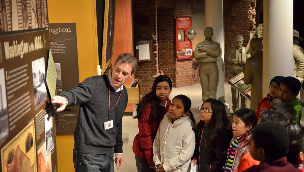 A man leads a group of children on a tour of the of a museum. He points to a picture on a display while the children watch.