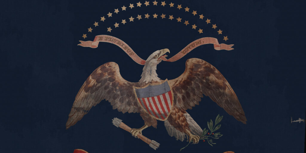 Photograph of a flag with the emblem of an eagle. The eagle is wearing a shield decorated as an American flag, and grasps a bundle of arrows and a spring of a plant in its talons. In its beak is a ribbon that reads "E. Pluribus Unum."