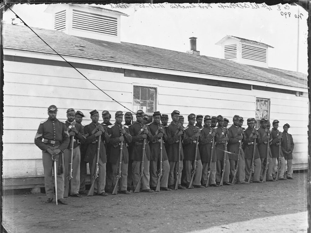 A row of Black men in Union army uniforms stand in front of a long white building.