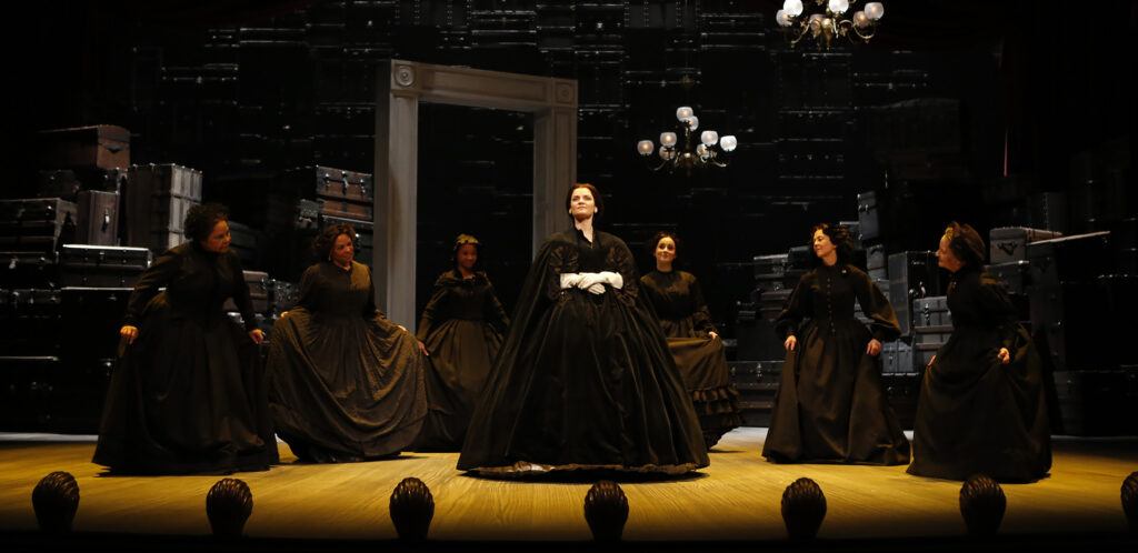 An actress portraying Mary Lincoln, dressed in a black gown and white gloves, kneels on stage. A Union solider in a blue uniform and yellow sash, with a gun behind his shoulder, looks at her. Behind them are two women wearing black.