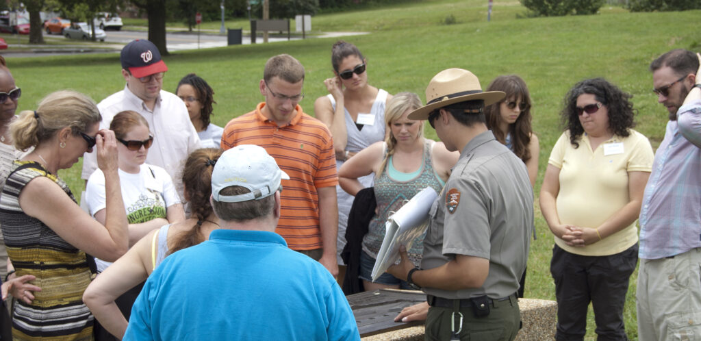 A group of men and women gather around a park ranger as he point something out on a memorial plaque.