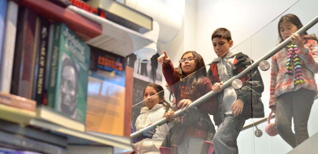 Young children stand behind and a railing and look at a tower of books.