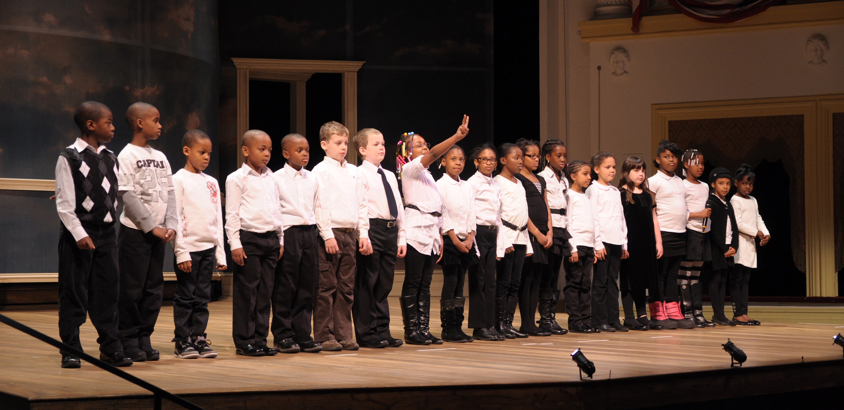 A row of children dressed mostly in white shirts and black pants stand in a line on a stage.