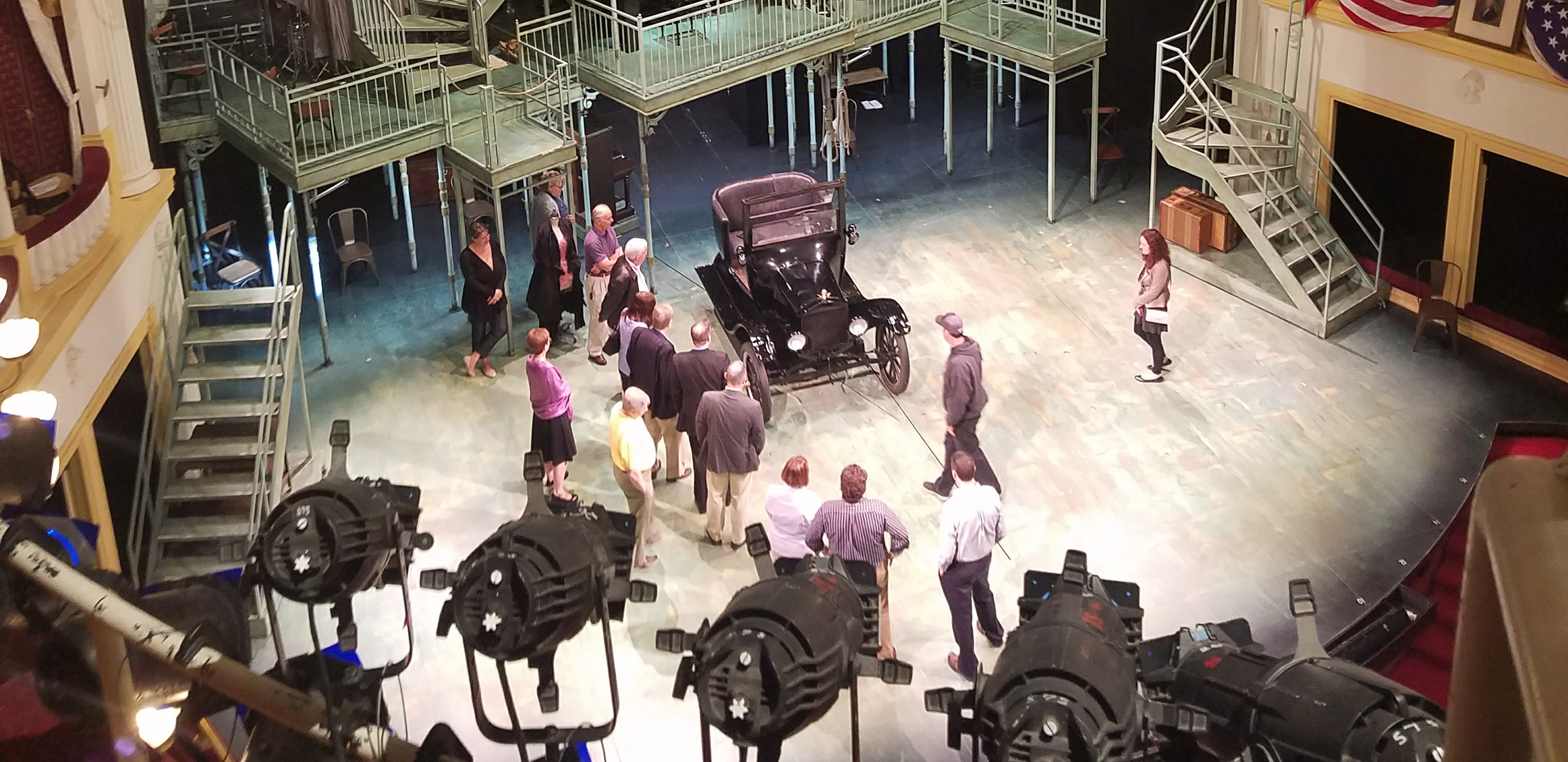 A group of people gather on a stage and examine a replica of an early automobile.