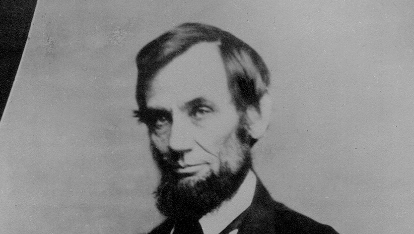 Abraham Lincoln, wearing his signature beard and dressed in a suit, is seated for a formal portrait. He is photographed from the waist up and looks directly into the camera.