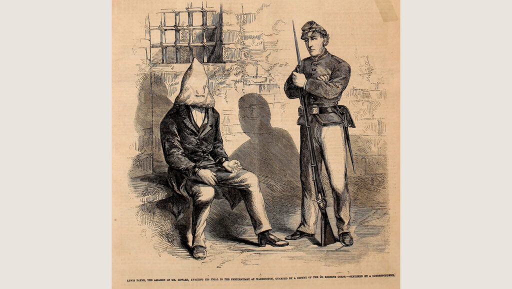 A union soldier guards a man in a hood and manacles sitting on a bench in a stone building.