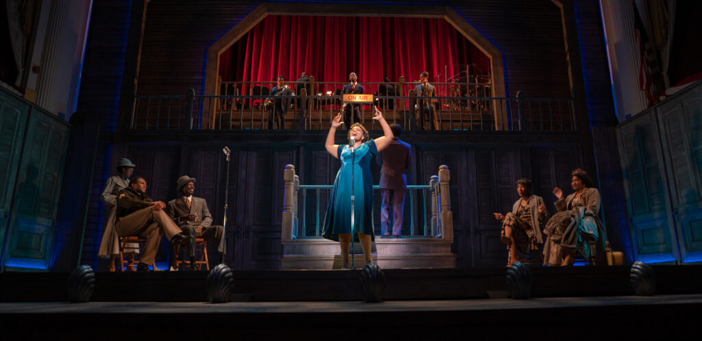 An African American woman wearing a two-toned teal dress stands center stage, singing in front of a microphone with an “on-air” sign behind her. Ensemble members look on from her left, right and on a platform above her. An onstage band is visible on a platform above her.