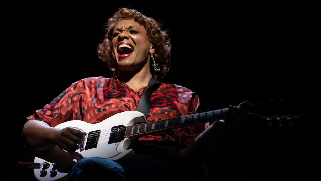 An African American woman wearing a red dress sits in a wheelchair center stage holding and playing a white electric guitar, singing with expression. 