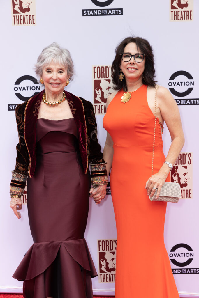 A woman wearing a maroon dress and jacket and a woman wearing an orange dress pose on a red carpet.