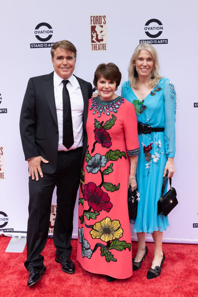 A man wearing a suit and tie, a woman wearing a pink dress with colorful floral accents and a woman wearing a light blue dress with smaller floral accents pose on a red carpet.