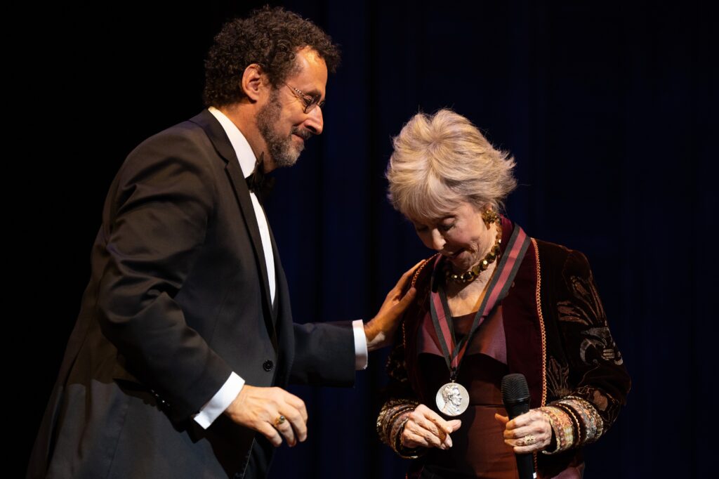 A man wearing a suit and glasses rests his hand on a woman’s shoulder as she looks down at her Lincoln Medal, which she is wearing around her neck.