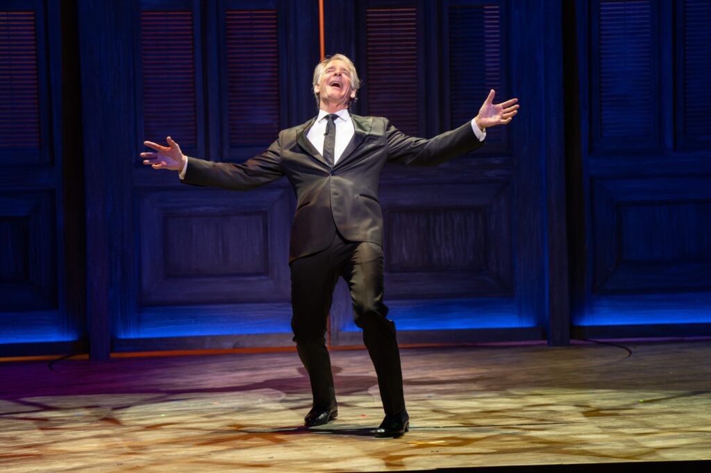 A man wearing a suit sings and dances on the Ford’s Theatre stage, looking up joyfully, with his arms outstretched.