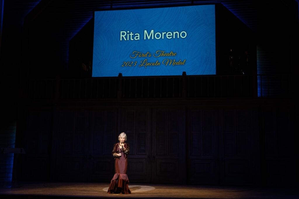 A woman wearing a maroon floor-length dress stands center stage. A PowerPoint slide is behind her, which reads “Rita Moreno. Ford’s Theatre 2023 Lincoln Medal.”