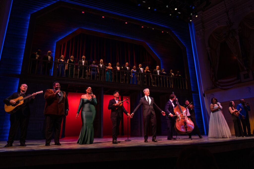A company of performers all dressed in gala wear assemble on the Ford’s Theatre stage. Featured talent stands on the Ford’s Theatre raked stage. The Ford’s Theatre ensemble stands on a balcony, with the house band behind them.