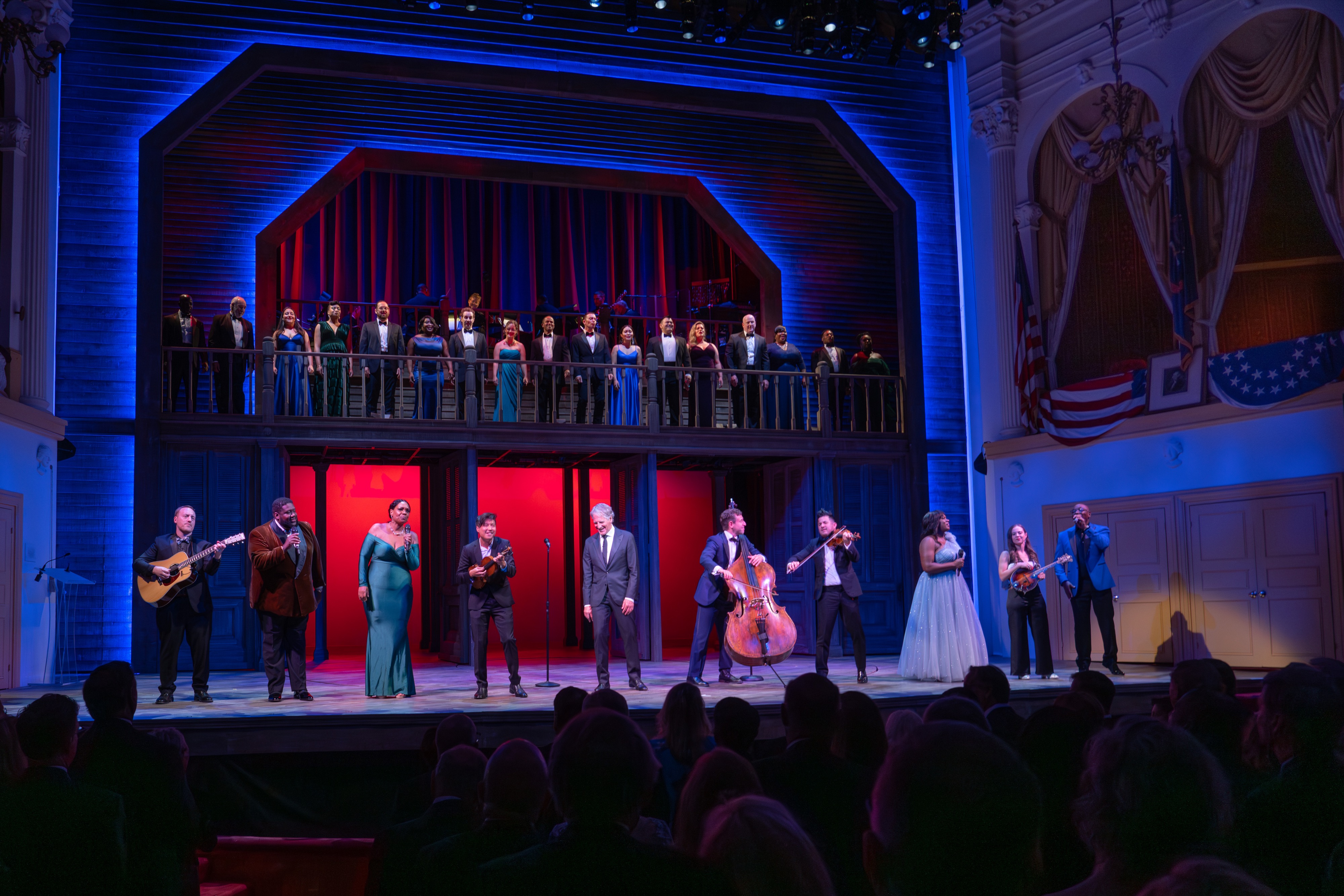A company of performers all dressed in gala wear assemble on the Ford’s Theatre stage. Featured talent stands on the Ford’s Theatre raked stage. The Ford’s Theatre ensemble stands on a balcony, with the house band behind them. The Presidential Box is visible on their right.