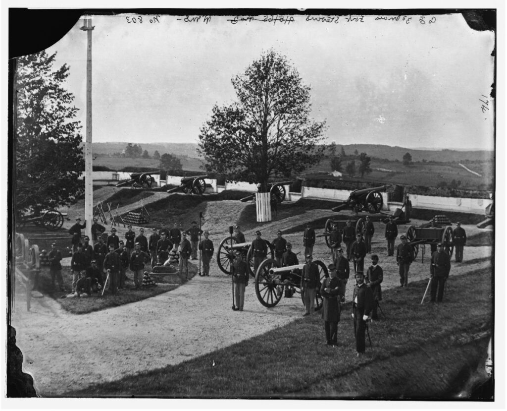 19th century photograph of Union soldiers dressed for battle, standing inside Fort Stevens with several cannons positioned, ready to fire. Most stand on the grass. 