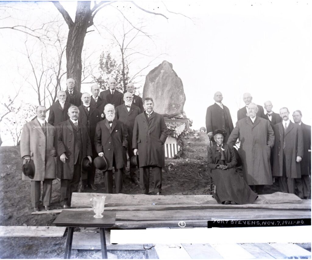 A group of elderly white veterans wearing suits stand next to a monument of President Abraham Lincoln, outside next to a tree. A few of them have hats in their hands. A Black woman wearing a long black skirt, jacket and hat sits on a chair with them. They all gaze with reverence into the camera.