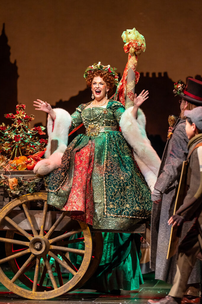 A white woman dressed as the Ghost of Christmas Present in a richly decorated green Victorian-style dress sits on a cart and smiles with a jovial expression, her arms outstretched.