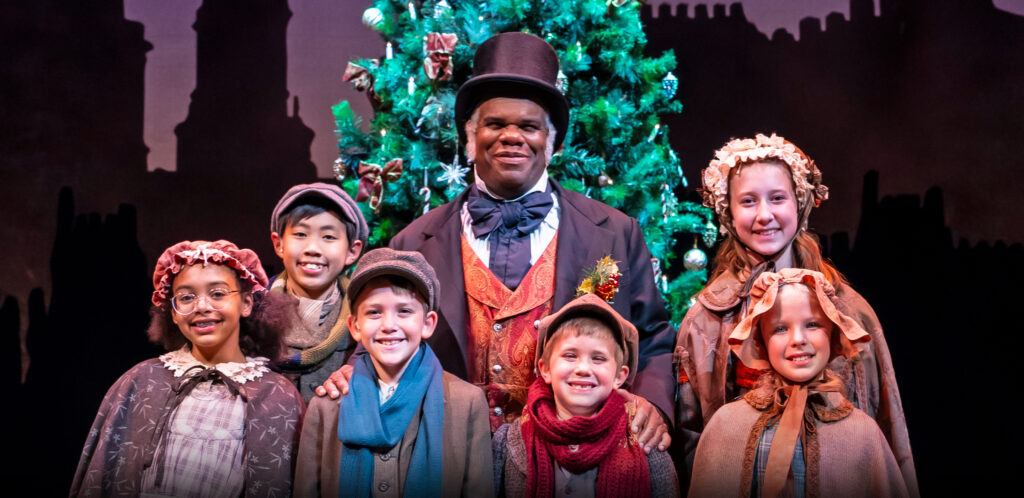 A smiling Black man in Victorian-style clothes and a top hat stands with a group of diverse children, also in Victorian-style clothing, in front of a large Christmas tree.