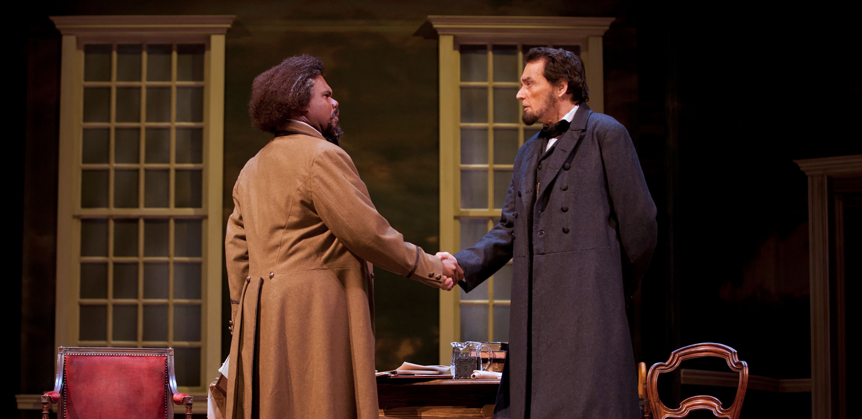 Actors portraying Frederick Douglass and Abraham Lincoln shake hands and talk while standing in front of a table and two closed windows.