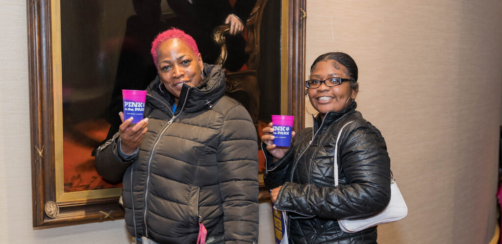 An older Black woman and a younger Black woman have fun at a post-show reception. They stand in front of a painting of Abraham Lincoln with drinks.