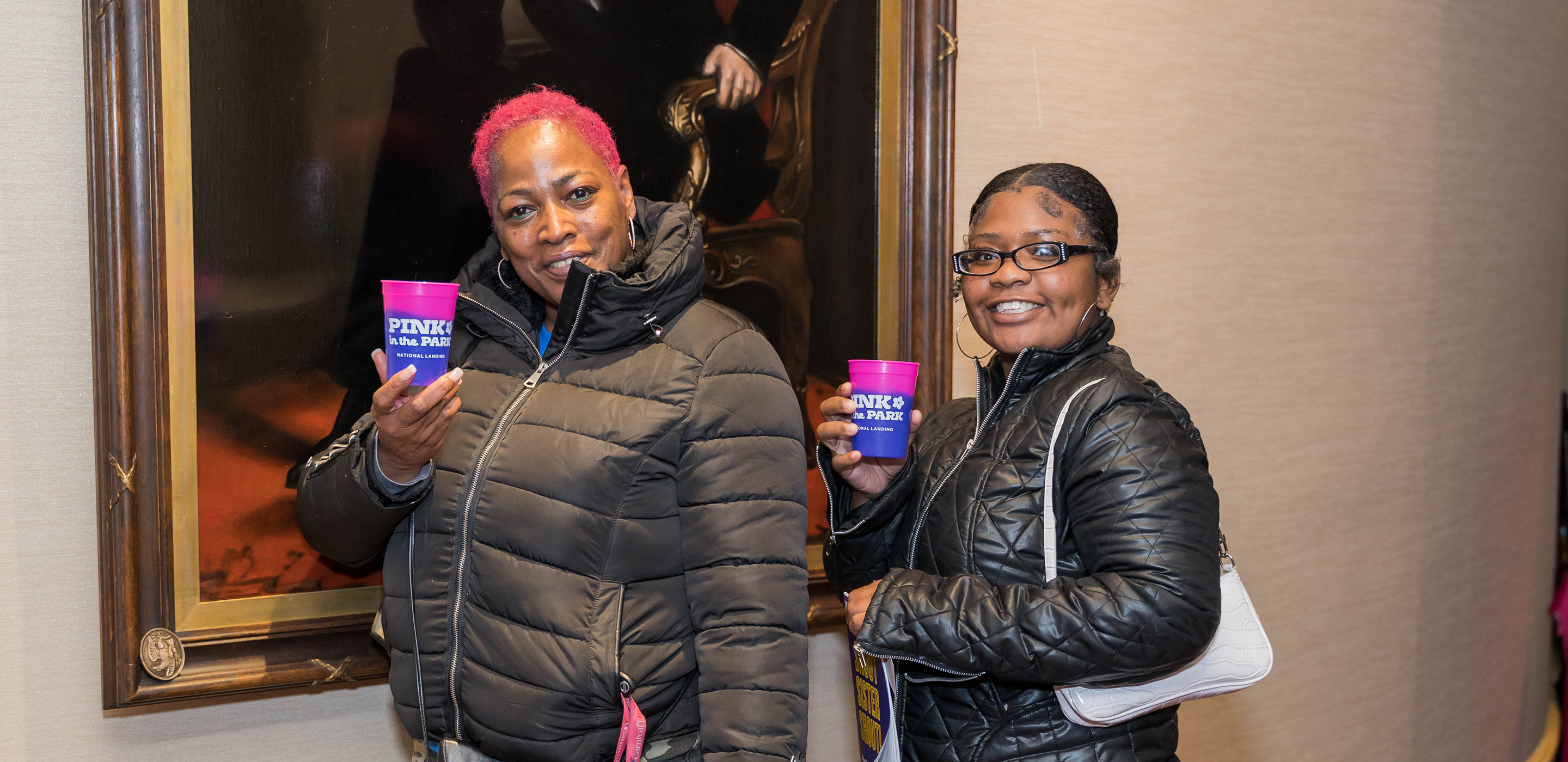 An older Black woman and a younger Black woman have fun at a post-show reception. They stand in front of a painting of Abraham Lincoln with drinks.