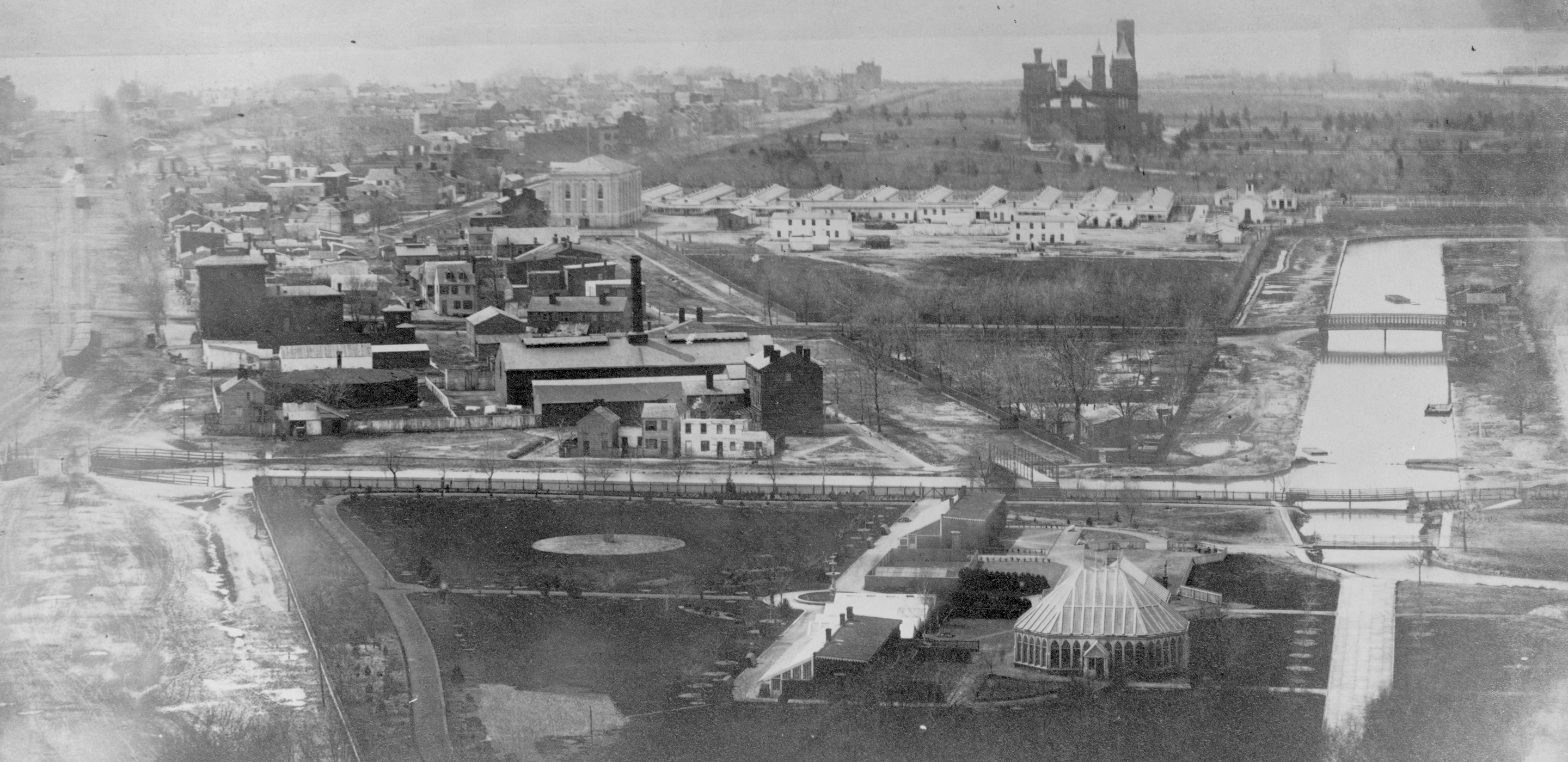 Black and white photograph overlooking the Washington City Canal; featuring muddy roads, a Civil War hospital, the Smithsonian Castle, and the Potomac River in the background.