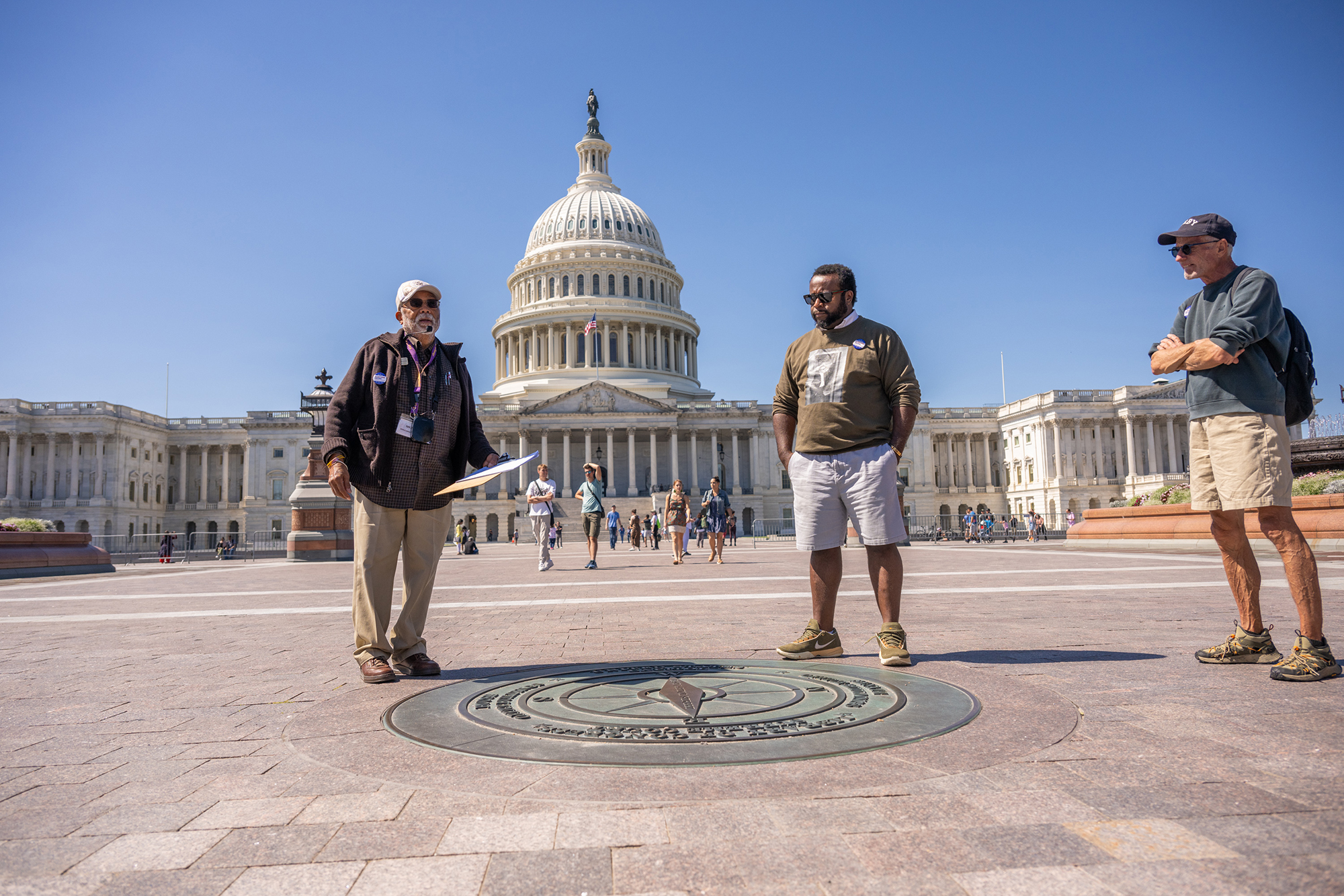Two tour guides stand next to a faded bronze compass on a plaza, in front of a historic building. A participant looks on. 