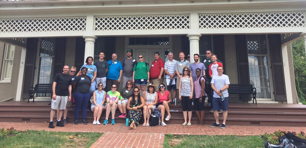 24 teachers arranged in two rows smile and pose for a group photo on the steps of Frederick Douglass National Historic site. Behind them is a large, Victorian house with a wide front porch with lattice trim, and large, floor to ceiling windows on either side of the front door.