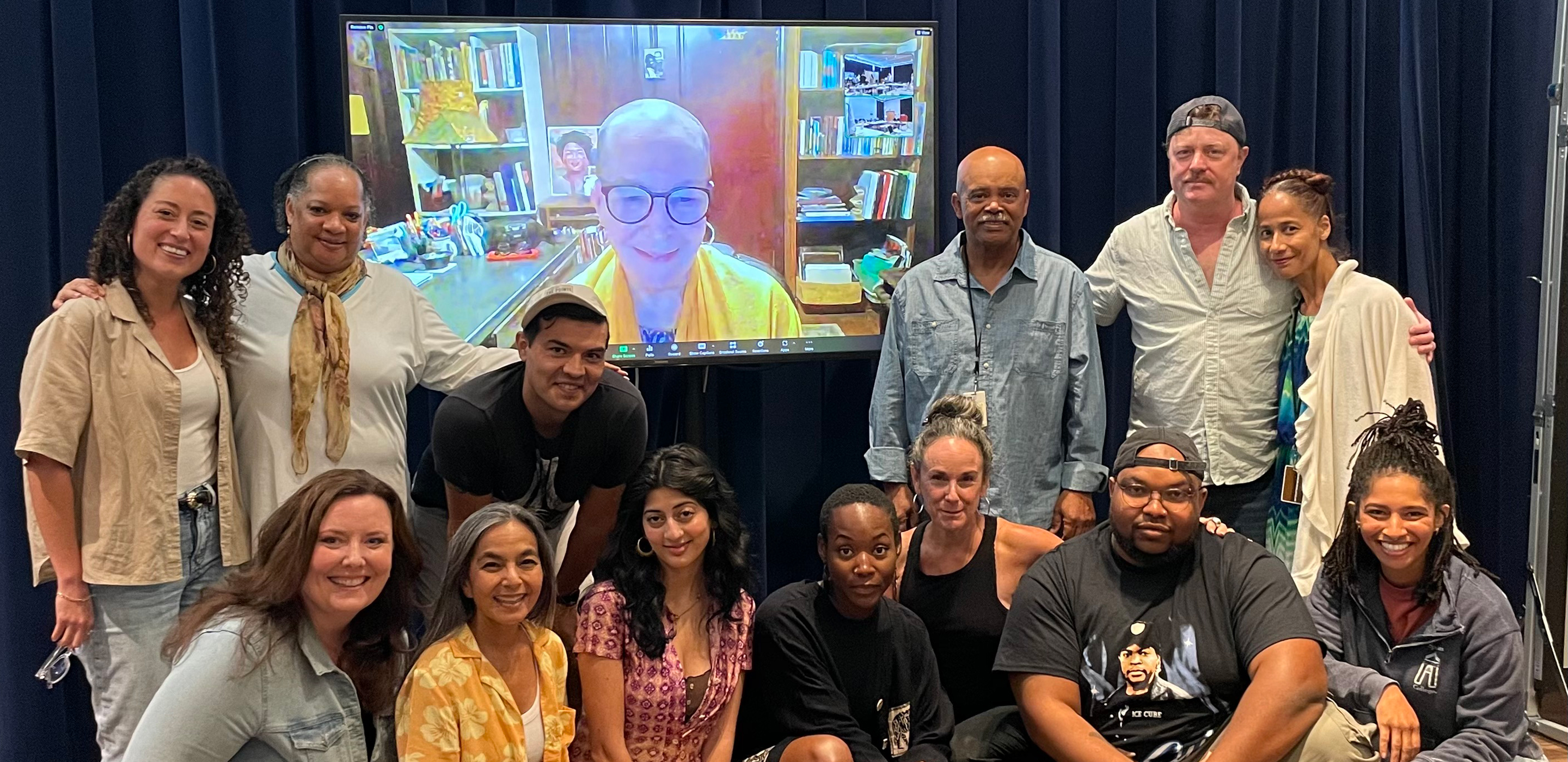 Thirteen people of various genders and ethnicities stand or sit around a large screen with a smiling woman on it.