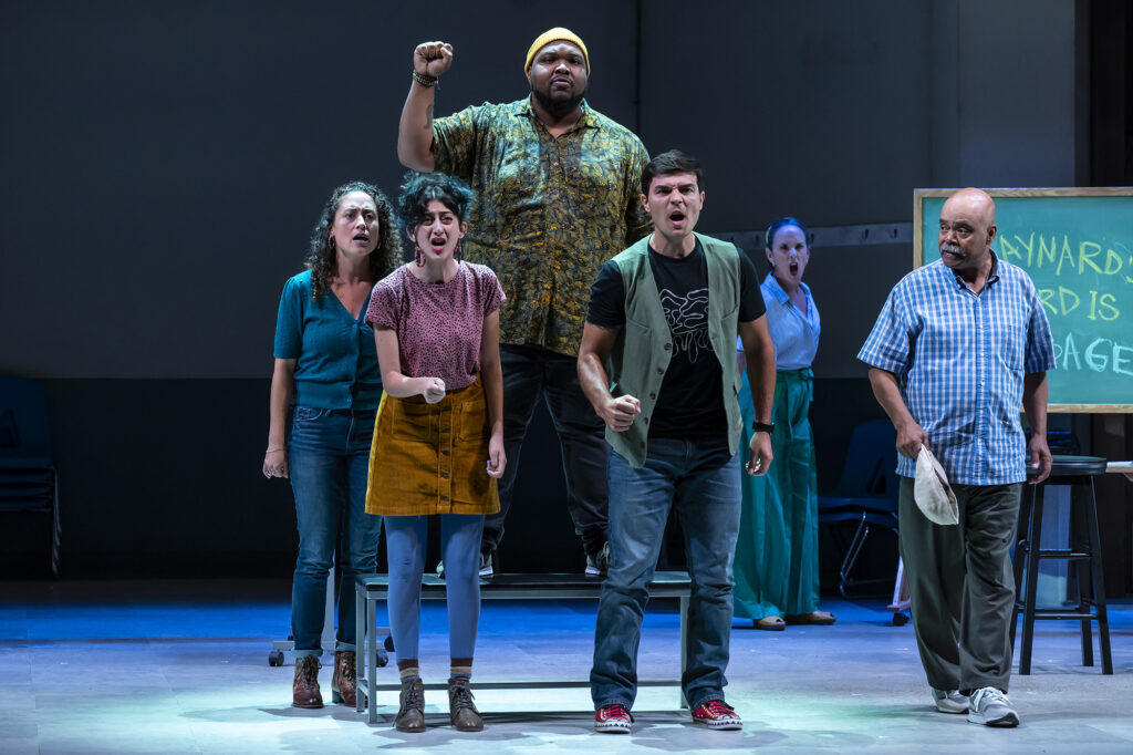 Six multicultural and intergenerational actors re-enact a union workers’ riot scene. One Black actor raises his fist with resolve. Next to them is a blackboard reading “Maynard’s Word Is Garbage.”