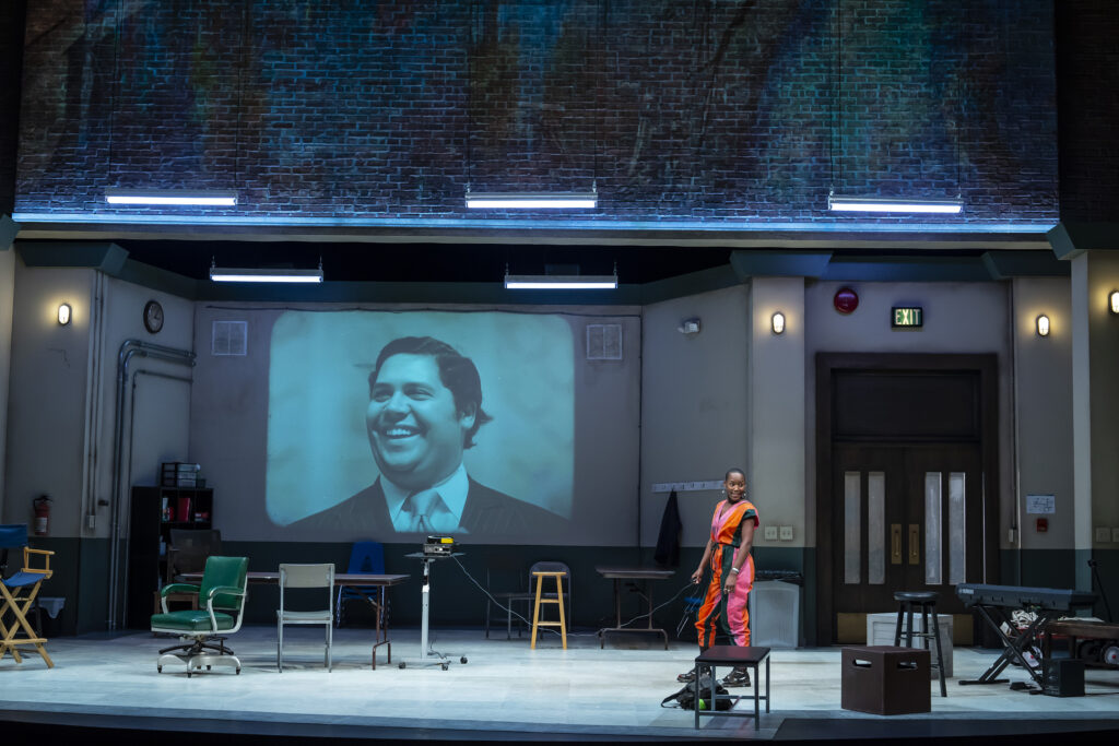 A Black actor wearing a brightly-colored jumpsuit stands center stage with a clicker and a projector, as a picture of Maynard Jackson is projected onto the wall of a rehearsal room. A brick wall is visible above the projection.